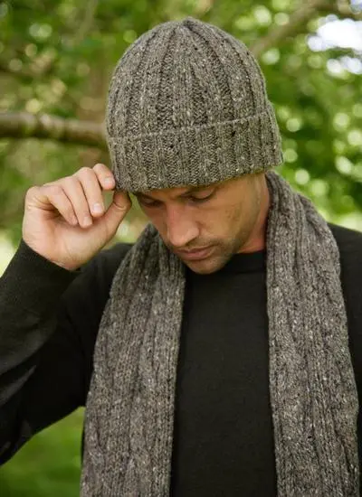 Close-up of man wearing a matching brown knit hat and scarf set looking down with greenery in background.